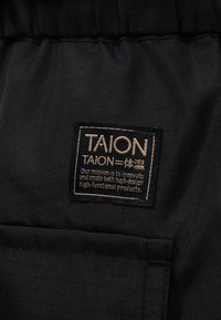 Μαύρο ύφασμα με ραμμένη ορθογώνια ετικέτα που αναγράφει "TAION" και μια δήλωση αποστολής σχετικά με την καινοτομία υψηλής σχεδίασης και υψηλής λειτουργικότητας προϊόντων.