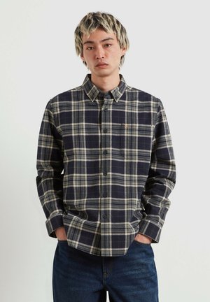 Giovane uomo con le mani in tasca che indossa una camicia a quadri nera e grigia con bottoni e jeans blu scuro su uno sfondo semplice.
