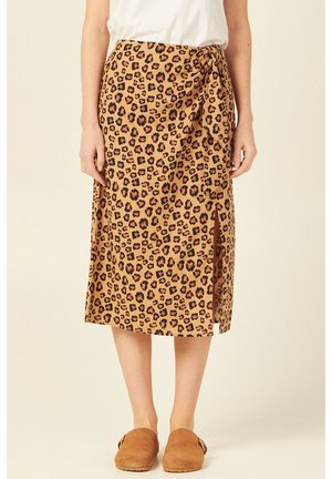 Femme portant une jupe beige imprimé léopard longueur genou avec une fente latérale et des chaussures marron sans lacets, debout devant un fond clair uni.
