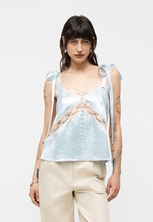 Donna con capelli scuri che indossa un camicino in raso azzurro chiaro con rifiniture in pizzo e spalline annodate, e pantaloni beige, su uno sfondo uniforme.