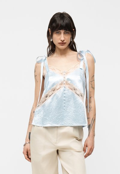Femme aux cheveux foncés portant un caraco en satin bleu clair orné de dentelle avec des bretelles nouées aux épaules et un pantalon beige, sur un fond uni.