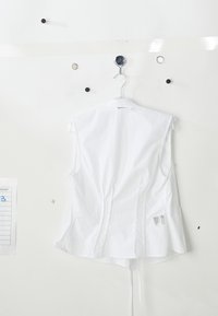 White sleeveless shirt hanging on a hook, featuring smooth fabric, a clean design, back seams, and a small tag on the lower side.