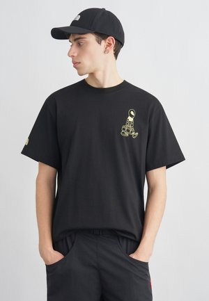 Young man in black cap, black t-shirt with small yellow astronaut graphic, and black pants, standing with hands in pockets, looking left.