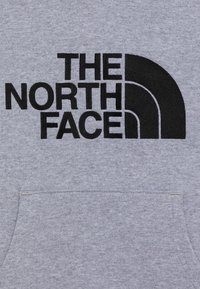 Felpa grigia con tasca frontale e logo stampato nero che presenta il testo "The North Face" e un design a mezz'arco. Tessuto morbido e strutturato.
