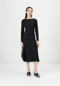 Robe noire à manches longues avec un corsage ajusté et un ourlet évasé, fabriquée en tissu doux. Portée avec des sandales à glisser noires et un petit sac à main.