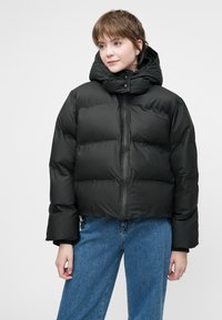 Tânără îmbrăcată într-o geacă puffer neagră cu guler înalt și glugă, purtată cu blugi din denim albastru pe un fundal simplu.
