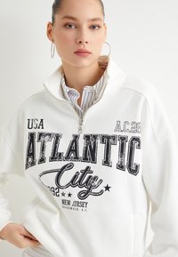 Sudadera blanca con cremallera que presenta gráficos negros con la inscripción "ATLANTIC CITY", con un cuello alto y mangas largas sobre una camisa de rayas.