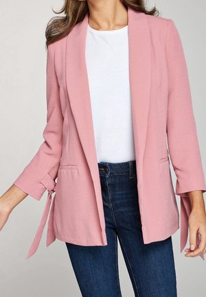 Mujer con una blazer abierta color rosa claro con puños anudados, una camiseta blanca de cuello redondo y vaqueros azul oscuro.