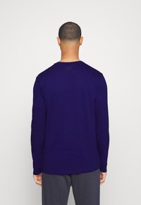 Man med kort, lockigt hår iförd en långärmad mörkblå Under Armour-tröja och mörkgrå byxor, vänd bort från kameran mot en enkel bakgrund.