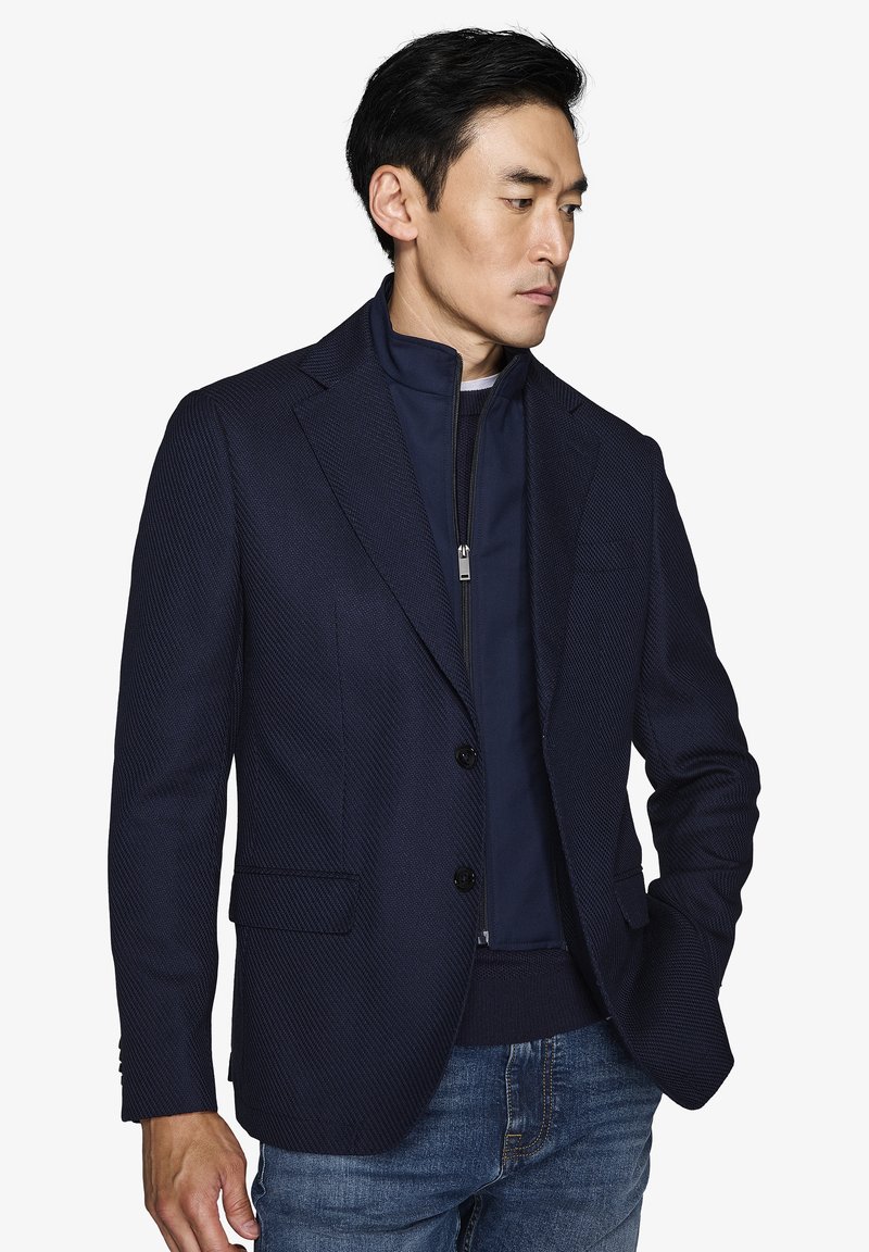 Skræddersyet mørkeblå blazer med subtil diagonal mønster, to frontlommer og to knapper. Båret over en navy lynlåsjakke.