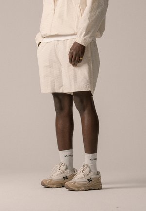 Vêtements de sport beige clair comprenant un haut zippé et un short assorti. Baskets beige avec accents noirs et chaussettes blanches épaisses.