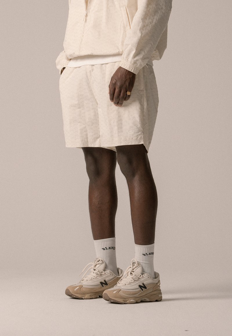 Light beige athletic apparel featuring a zip-up top and matching shorts. Sneakers are beige with black accents and thick white socks.