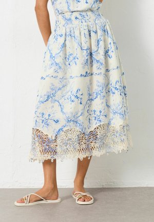 Frau trägt einen weißen Midirock mit blauem Blumenmuster und Spitzenbesatz, kombiniert mit weißen flachen Riemchensandalen auf einem grauen Boden.