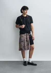 Zwart poloshirt met rode accenten, zwarte short en een geruite wikkelrok. Zwarte schoenen en witte sokken maken de outfit compleet. Casual stijl.