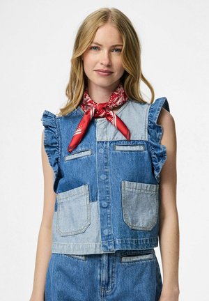 Femme aux cheveux blonds portant un gilet en jean patchwork sans manches et un jean assorti, avec un foulard rouge à motif noué à l'avant.