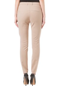 Nainen, joka pukeutuu beigekuituiset slim-fit-housut ja mustat korkokengät, seisoo selkä kameraan päin valkoista taustaa vasten.