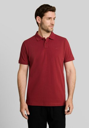 Man die een effen bordeaux rood korte mouw polo draagt en een zwarte broek, kijkt opzij tegen een effen lichte achtergrond.