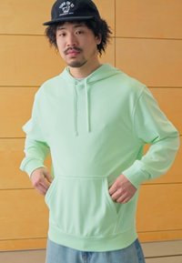 Sweatshirt verde menta com um bolso canguru à frente, capuz com cordão, ajuste solto e punhos canelados, combinado com jeans azuis claros.