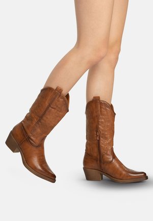 Beine, die braune Cowboy-Stiefel aus Leder tragen, die bis zur Mitte der Wade reichen, mit niedrigen, gestapelten Absätzen und Zickzack-Nähmustern auf weißem Hintergrund.