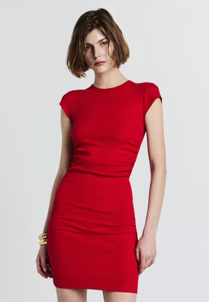 Donna con capelli corti castani che indossa un abito rosso aderente a coste e braccialetti d'oro, in piedi davanti a uno sfondo chiaro e uniforme.