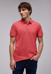 Polo shirt en coton corail à manches courtes, avec un plastron à trois boutons et un petit logo sur la poitrine ; associé à un jean bleu foncé.