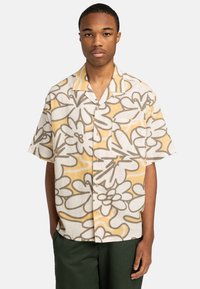 Joven de frente que lleva una camisa de bot�ón de manga corta con grandes patrones florales en beige y amarillo, y pantalones verde oscuro.