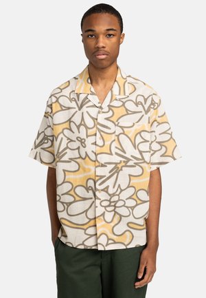 Young man facing forward wearing a short-sleeve button-up shirt with large beige and yellow floral patterns and dark green pants.