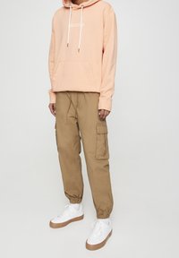 Felpa pesca con coulisse, tasca a marsupio; pantaloni cargo beige con tasche laterali; sneakers bianche con suola marrone.
