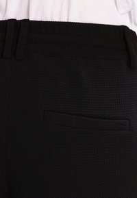 Close-up van zwarte gestructureerde broek met een tailleband en een enkele achterzakdetail tegen een witte stofachtergrond.