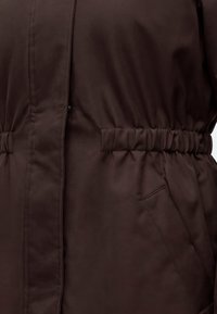 Manteau en tissu marron avec taille élastique et poche latérale avec coutures visibles.