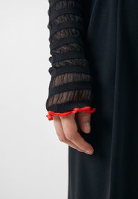 Main dans une manche rayée noire transparente avec un poignet rouge à volants, reposant contre un fond de vêtement noir.
