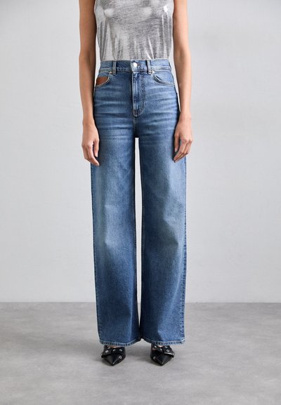 Jean en denim bleu à jambes larges avec une taille haute, présentant un design classique à cinq poches et un patch en cuir marron contrastant.