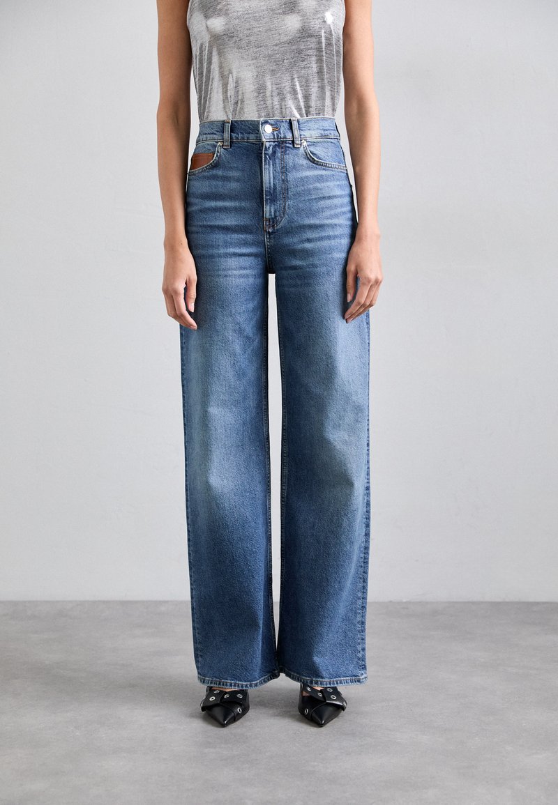 Pantalones vaqueros de mezclilla azul de pierna ancha con una cintura alta, que presentan un diseño clásico de cinco bolsillos y un parche de cuero marrón en contraste.