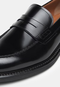 Mocassins en cuir noir avec une finition lisse, comportant une sangle décorative et un subtil détail de couture le long du bord et de la semelle.