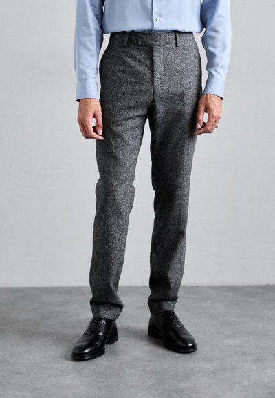 Mężczyzna ubrany w szare wełniane spodnie, czarne eleganckie buty oraz jasnoniebieską koszulę zapinaną na guziki, stojący na szarej podłodze wzdłuż gładkiej ściany.
