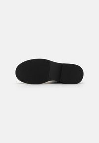 Semelle en caoutchouc noir avec une surface texturée et striée. La forme est plate avec un contour lisse et un point de connexion central.