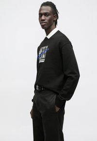 Sudadera negra con cuello blanco, presenta un estampado gráfico en negro y azul. Detalles incluyen un diseño ajustado y acentos visibles de cinturón.