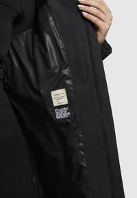 Interiorul jachetei negre are o textură netedă și lucioasă. Etichetele afișează instrucțiuni de îngrijire și numele brandului. Detaliul buzunarului pliat este vizibil.