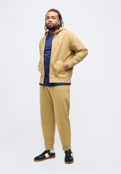 Άνδρας με ντραντλοκς, φορώντας ταμπά φόρμα και μπλε πουκάμισο, στέκεται με τα χέρια στις τσέπες σε απλό φωτεινό φόντο.