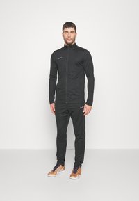 Sort atletisk træningsdragt med en zip-jakke og matchende bukser. Har Nike-logo i hvid. Stoffet ser glat og tætsiddende ud.