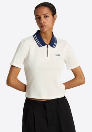 Femme portant un haut blanc à manches courtes avec col bleu marine et logo Vans, associé à un pantalon noir taille haute, debout les mains dans le dos.