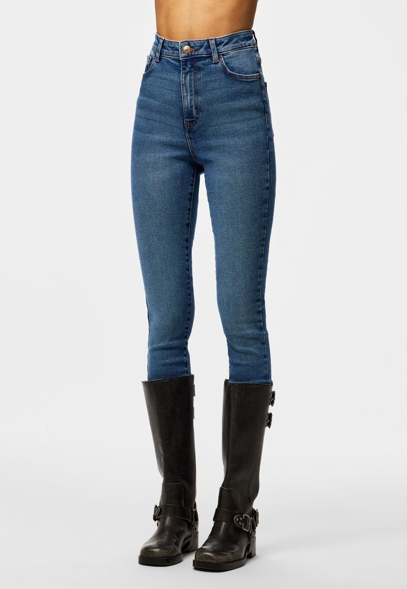 Personne portant un jean skinny bleu taille haute et des bottes en cuir noires montant jusqu'aux genoux avec des boucles, se tenant contre un fond blanc uni.