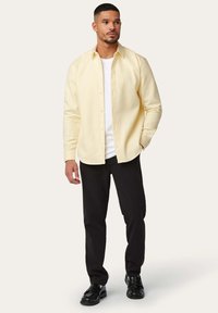 Gelbes Hemd mit Knopfleiste über einem weißen T-Shirt, kombiniert mit schwarzen Hosen und schwarzen Schuhen. Das Hemd hat einen lässigen Schnitt und lange Ärmel.