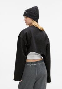 Črna skrajšana svetrovka z nizkimi rameni, mehka tekstura, v kombinaciji z oprijetim belim topom in sivimi hlačami z elastičnim pasom. Črna rebrasta kapa na glavi.