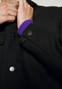 Mani che regolano il colletto di una giacca nera con bottoni metallici sopra un polsino di maglione viola.