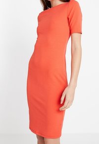 Apelsinfärgad klänning med rund halsringning och korta ärmar, tillverkad av textil med struktur, som slutar strax nedanför knäna. Inga synliga knäppningar.