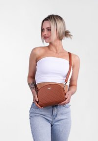 Hnědá syntetická crossbody taška s perforovaným designem, kulatým tvarem, nastavitelným popruhem a logem na přední straně.