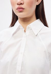 Λευκό πουκάμισο �με κουμπιά, με δομημένο γιακά και λεπτή γυαλάδα. Διαθέτει εφαρμοστή γραμμή, λεία υφή και διαφανή πλαστικά κουμπιά.