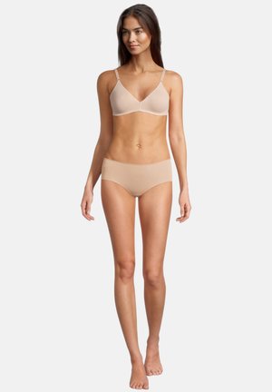 Femme debout pieds nus portant un soutien-gorge beige sans coutures et un slip hipster assorti, sur un fond clair uni.