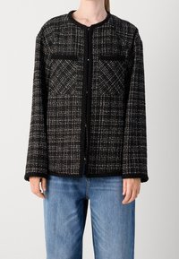 Femme portant une veste texturée à carreaux noir et blanc avec un col rond et un jean en denim bleu, debout devant un fond uni.